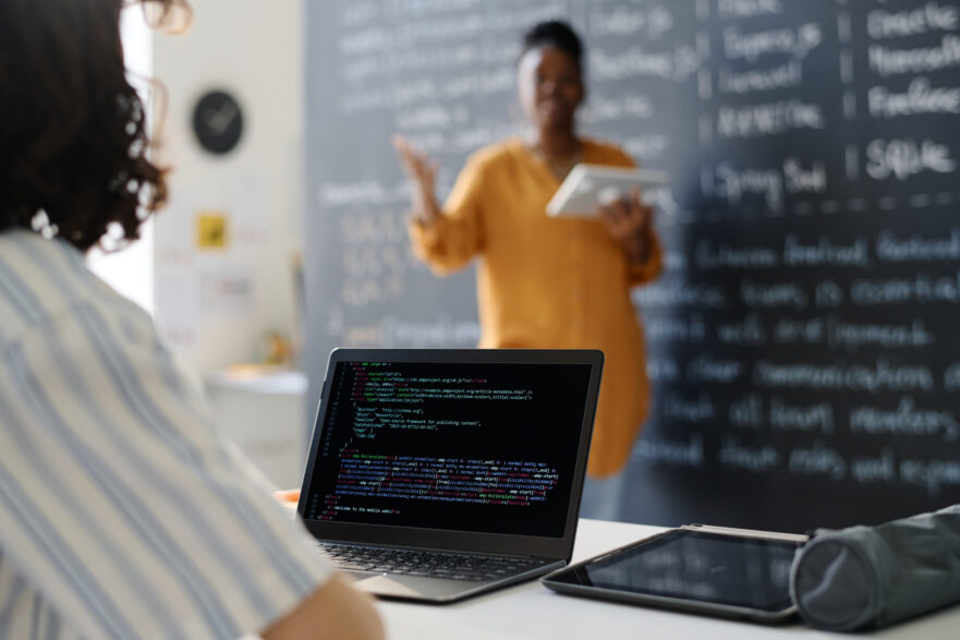 Vista posteriore dello studente che digita i codici sul computer mentre è seduto alla scrivania a lezione con l'insegnante che spiega il materiale in background (Depositphotos)